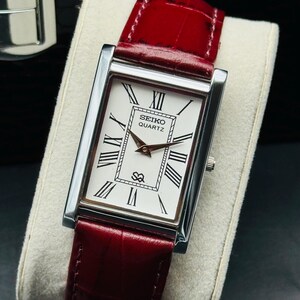 May include: A rectangular Seiko Quartz watch with a silver case and a white face featuring black Roman numerals. The watch has a red leather band. The watch is displayed on a light-colored surface.