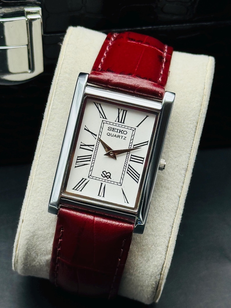 May include: A rectangular Seiko Quartz watch with a silver case and a white face featuring black Roman numerals. The watch has a red leather band. The watch is displayed on a light-colored surface.