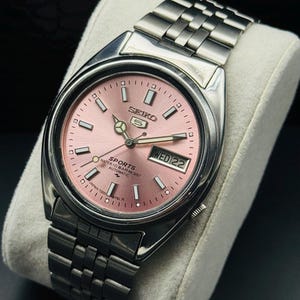 May include: A stainless steel Seiko 5 Sports automatic watch with a pink dial. The watch features a day and date display at the 3 o'clock position and a silver link bracelet. The watch is displayed on a white cushion.