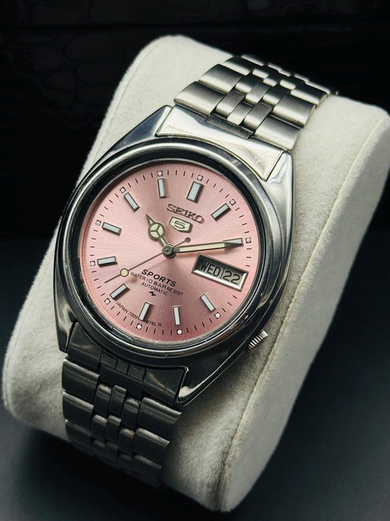May include: A stainless steel Seiko 5 Sports automatic watch with a pink dial. The watch features a day and date display at the 3 o'clock position and a silver link bracelet. The watch is displayed on a white cushion.