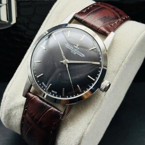 May include: A silver-toned Jaeger-LeCoultre Club watch with a dark gray face and a brown leather band. The watch has silver hands and markers, and the band is textured. The watch is displayed on a white cushion.