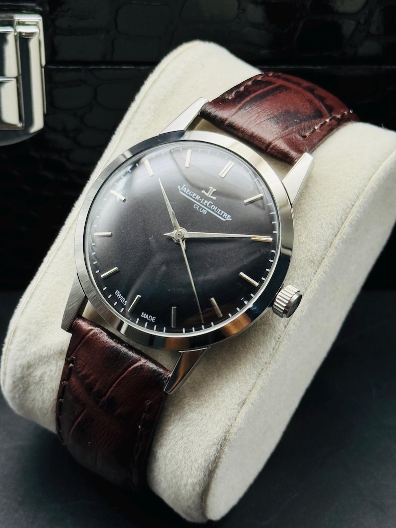 May include: A silver-toned Jaeger-LeCoultre Club watch with a dark gray face and a brown leather band. The watch has silver hands and markers, and the band is textured. The watch is displayed on a white cushion.
