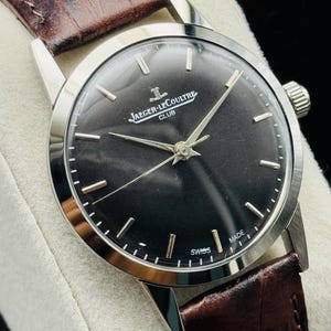 May include: A silver-toned Jaeger-LeCoultre Club watch with a dark face and brown leather band. The watch face displays the brand name and the word "SWISS MADE". The band is a rich brown color.