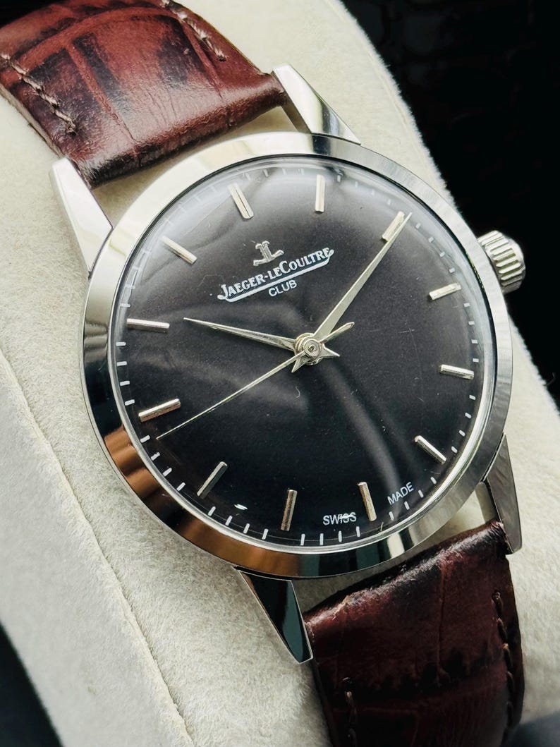 May include: A silver-toned Jaeger-LeCoultre Club watch with a dark face and brown leather band. The watch face displays the brand name and the word "SWISS MADE". The band is a rich brown color.