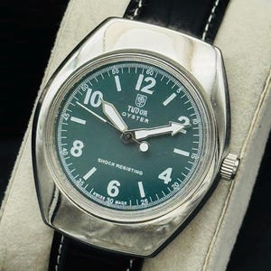 May include: A silver Tudor Oyster watch with a dark green face and black leather strap. The watch face features white numerals and hands, with the text "TUDOR OYSTER" and "SHOCK RESISTING". The strap is stitched with white thread.