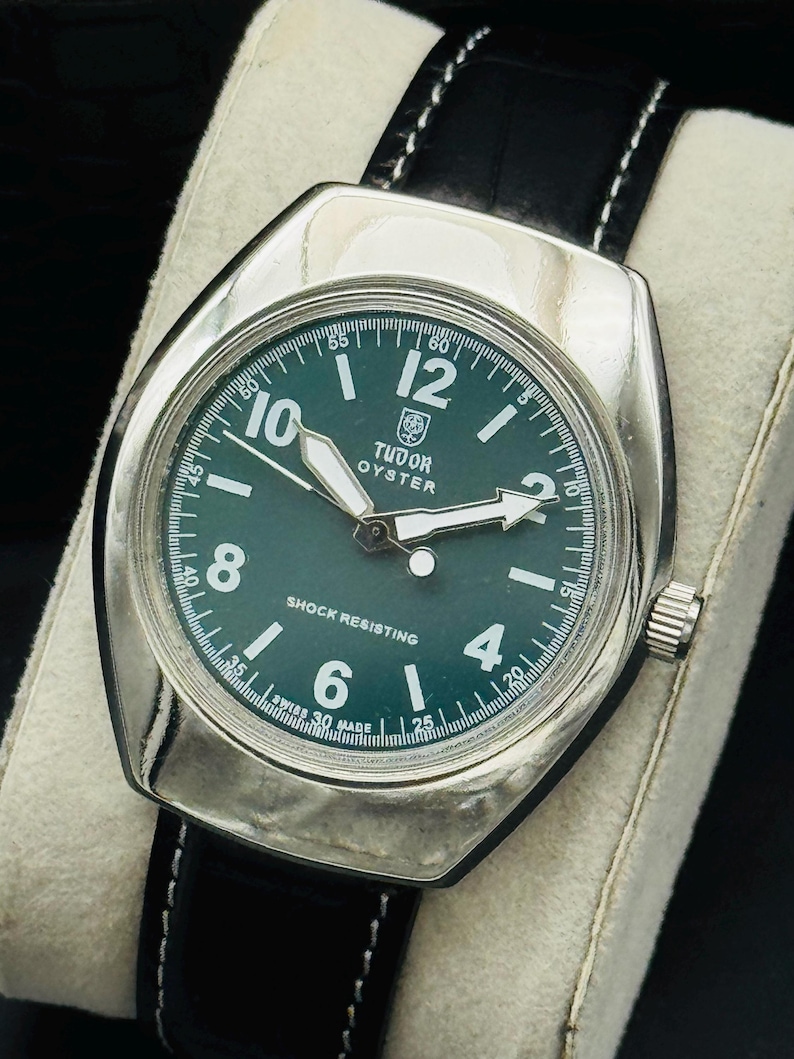 May include: A silver Tudor Oyster watch with a dark green face and black leather strap. The watch face features white numerals and hands, with the text "TUDOR OYSTER" and "SHOCK RESISTING". The strap is stitched with white thread.