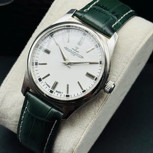 May include: A silver wristwatch with a white face and black markers. The watch has a green leather band with white stitching. The watch face has the text "JAEGER-LECOULTRE CLUB SWISS MADE T". The watch is displayed on a gray pillow.