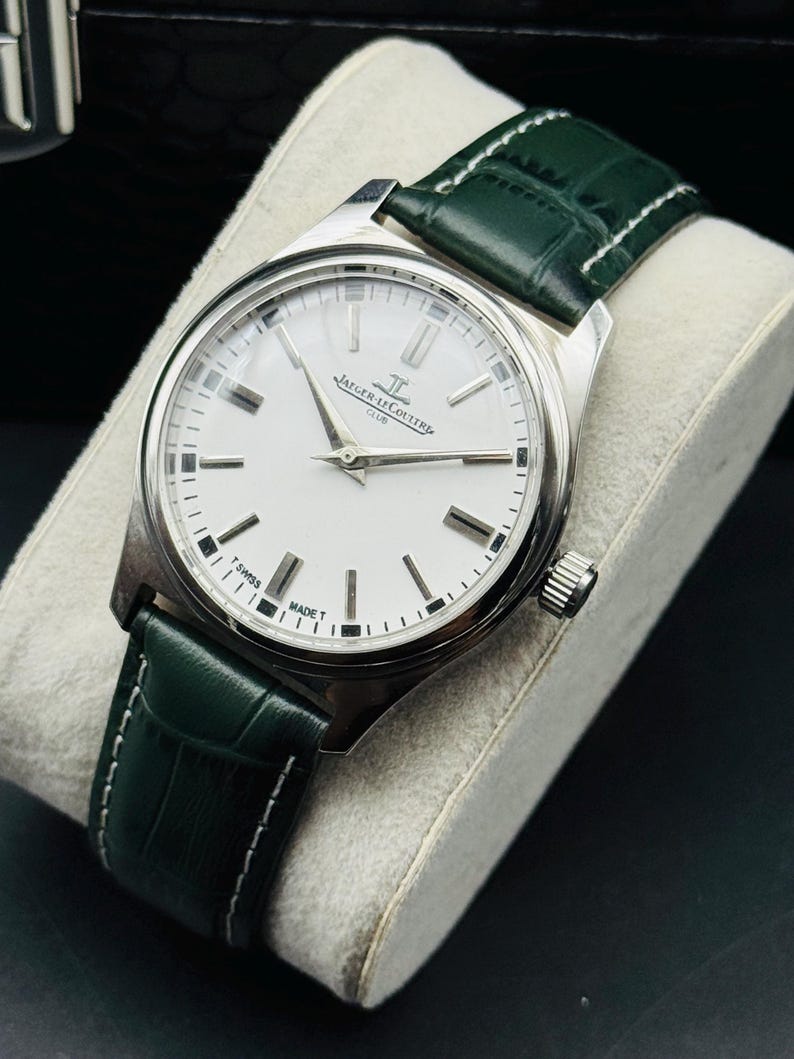 May include: A silver wristwatch with a white face and black markers. The watch has a green leather band with white stitching. The watch face has the text "JAEGER-LECOULTRE CLUB SWISS MADE T". The watch is displayed on a gray pillow.