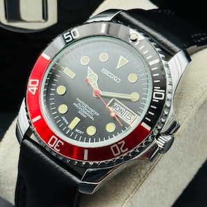May include: A stainless steel Seiko Marine Master automatic watch with a black dial, red bezel, and black leather strap. The watch face displays the day and date. The watch is water resistant to 300m.