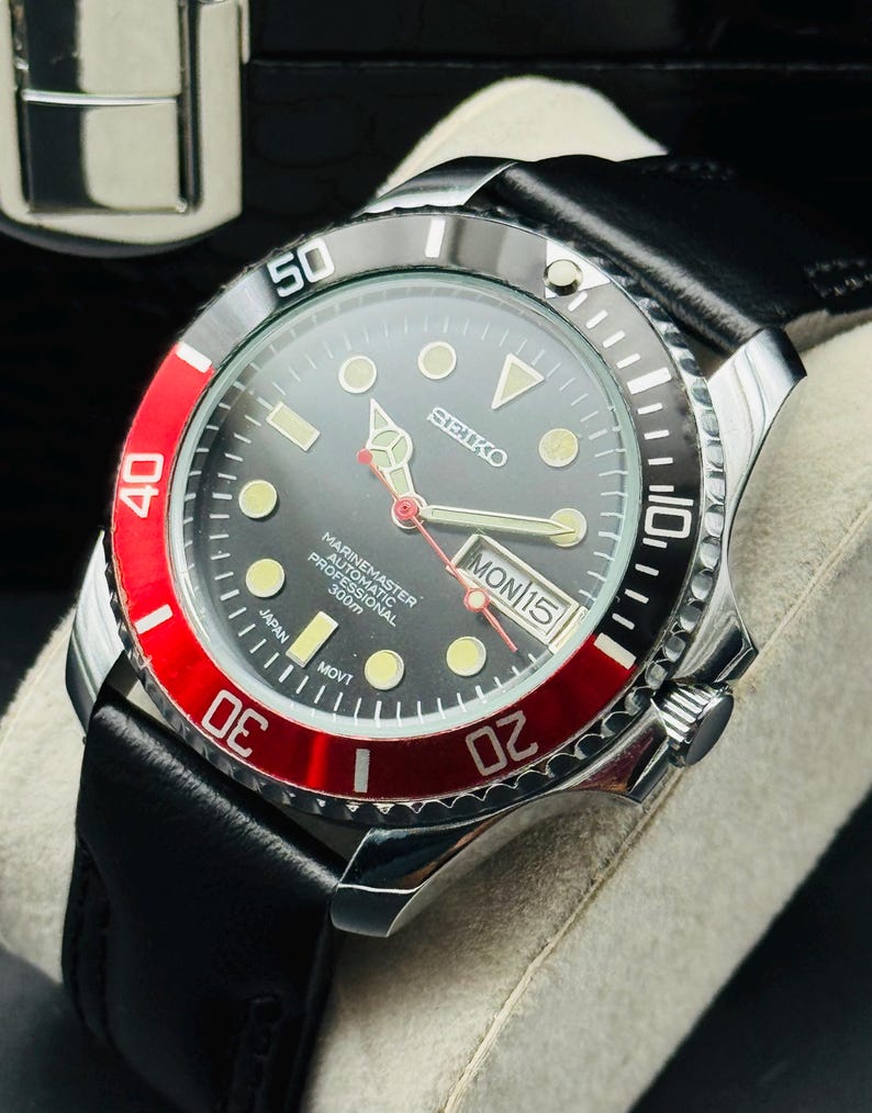 May include: A stainless steel Seiko Marine Master automatic watch with a black dial, red bezel, and black leather strap. The watch face displays the day and date. The watch is water resistant to 300m.