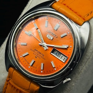 Puede incluir: Un reloj automático Seiko 5 Sports vintage con esfera naranja y caja de acero inoxidable. El reloj tiene una pantalla de día y fecha y una correa de cuero naranja. La esfera incluye el texto "Seiko 5 Sports Water 10 Bar Resist Automatic".