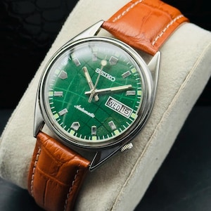 May include: A vintage Seiko automatic wristwatch with a green dial and a brown leather strap. The watch face displays the day and date. The watch is set on a cream-colored cushion.