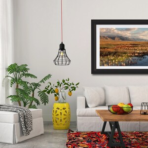 May include: A framed landscape photograph depicting a serene marsh scene with mountains in the background. The artwork is displayed in a modern living room setting, alongside a white sofa, a wooden coffee table, and decorative plants.