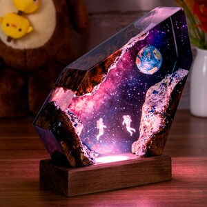Peut inclure: Lampe décorative représentant une scène spatiale, enfermée dans de la résine transparente. La lampe représente des astronautes, une planète et une galaxie étoilée. La base est en bois. La lampe émet une douce lueur chaude.