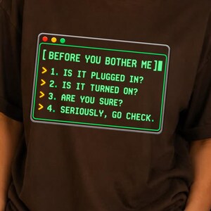 Peut inclure: Un t-shirt à manches courtes marron foncé avec un graphique représentant une fenêtre de terminal informatique et du texte vert : [ BEFORE YOU BOTHER ME ] > 1. IS IT PLUGGED IN? > 2. IS IT TURNED ON? > 3. ARE YOU SURE? > 4. SERIOUSLY, GO CHECK.