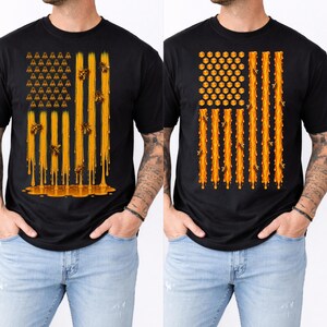 Può includere: Una t-shirt nera con due distinti disegni grafici. Il disegno a sinistra mostra un motivo a bandiera fatto di gocce di miele e api. Il disegno a destra mostra un motivo a bandiera composto da celle esagonali e gocce di miele con api.