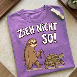 May include: A heather purple t-shirt featuring a graphic of a brown sloth walking a turtle on a leash. The text "ZIEH NICHT SO!" is printed in white above the sloth. The sloth has a tired expression, and the turtle wears a blue collar.