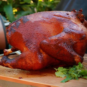 May include: A whole roasted turkey with a deep reddish-brown, crispy skin texture. The cooked poultry is positioned on a wooden surface, accompanied by a small cluster of fresh green cilantro leaves placed near the base of the bird.
