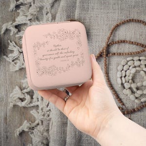 May include: A light pink jewelry box with a floral design and the text "Rather, it should be that of your inner self, the unfading beauty of a gentle and quiet spirit 1 PETER 3:4". The box is held in a hand.