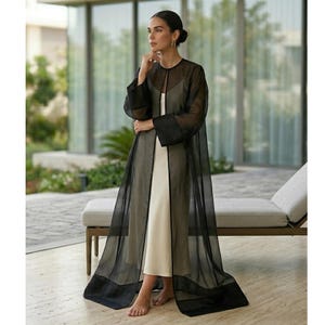 Può includere: Una donna indossa un soprabito nero trasparente sopra un vestito color crema. Il soprabito è lungo, fluente e ha maniche ampie. L'abito è un design semplice ed elegante. L'outfit è adatto alle occasioni formali.