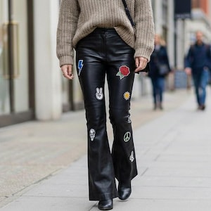 Könnte beinhalten: Schwarze Schlaghose aus Leder, verziert mit bunten Aufnähern, darunter ein Blitz, ein Friedenszeichen und eine Rose. Die Hose hat eine hohe Taille und einen weiten Beinabschluss, was einen Retro-Stil erzeugt.