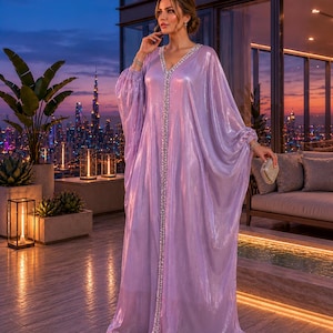 Puede incluir: Un vestido largo y fluido de color lavanda con escote en V y adornos brillantes. El vestido presenta mangas anchas y se usa con zapatos dorados. El fondo muestra un horizonte urbano al anochecer.