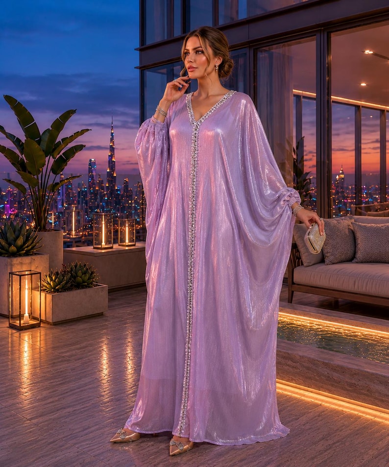 Puede incluir: Un vestido largo y fluido de color lavanda con escote en V y adornos brillantes. El vestido presenta mangas anchas y se usa con zapatos dorados. El fondo muestra un horizonte urbano al anochecer.