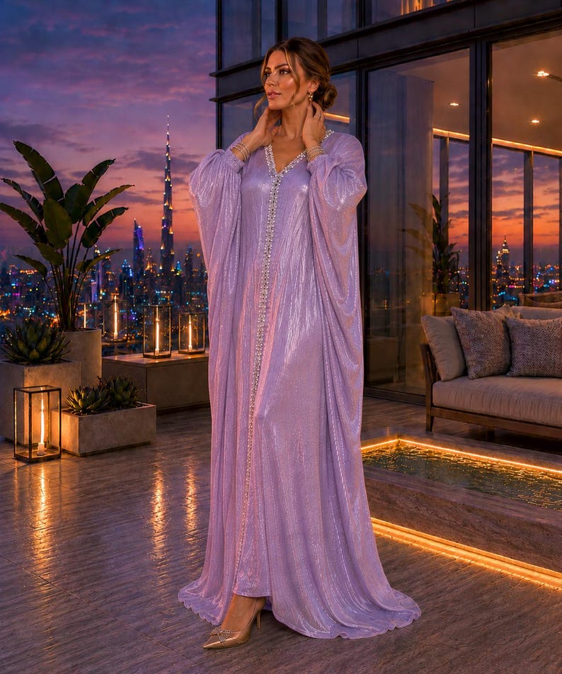 Puede incluir: Un vestido largo y fluido de color lavanda con escote en V y adornado con una l&iacute;nea de piedras brillantes en la parte delantera. El vestido tiene mangas largas y drapeadas y se usa con tacones dorados. El fondo presenta un horizonte urbano al atardecer.