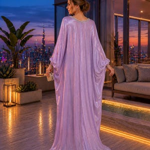 Puede incluir: Un vestido caft&aacute;n brillante de color lavanda con un escote con cuentas. El vestido tiene mangas largas y fluidas y un dobladillo hasta el suelo. La mujer est&aacute; de pie en un balc&oacute;n con vistas a un horizonte urbano al anochecer, sosteniendo un peque&ntilde;o bolso dorado.