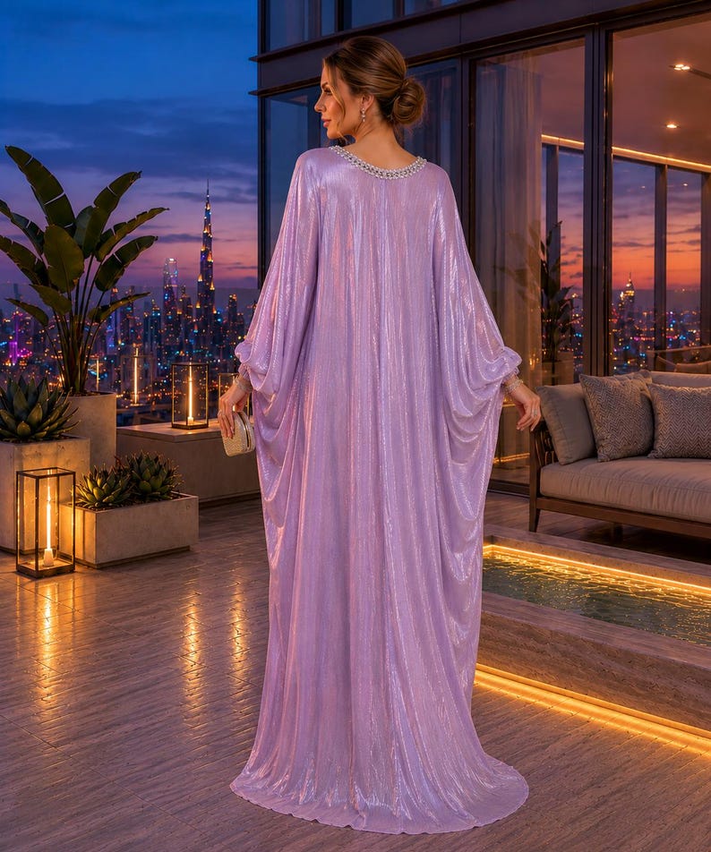 Puede incluir: Un vestido caft&aacute;n brillante de color lavanda con un escote con cuentas. El vestido tiene mangas largas y fluidas y un dobladillo hasta el suelo. La mujer est&aacute; de pie en un balc&oacute;n con vistas a un horizonte urbano al anochecer, sosteniendo un peque&ntilde;o bolso dorado.
