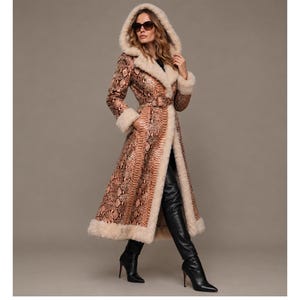 May include: A long, hooded coat featuring a brown and tan snakeskin pattern, accented with thick cream-colored faux fur trim along the hood, cuffs, and hem. The coat includes a matching belt at the waist and is paired with black leather high-heeled boots.