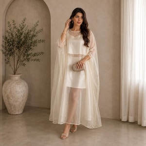 Puede incluir: Un conjunto de color crema hasta el suelo que presenta un vestido interior sin mangas con escote redondo y un caftán superpuesto transparente, ligero y de manga larga. El atuendo se combina con un pequeño bolso de mano rectangular a juego.