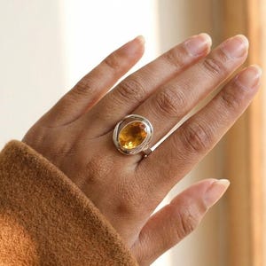 May include: A silver ring featuring a large, oval-cut, golden-yellow gemstone. The gemstone is set in a bezel setting with a swirling design around it. The ring is shown on a hand with a brown sleeve in the background.