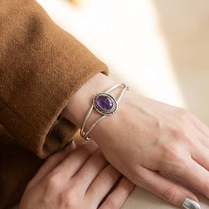 Può includere: Un bracciale a polsino in argento con una gemma ovale viola. La gemma è circondata da un bordo di perline. Il bracciale è indossato su un polso, con una manica di giacca marrone sullo sfondo.