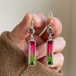 May include: A pair of silver earrings with a hook closure. Each earring features a small, round, multi-colored stone at the top, and a larger, rectangular, multi-colored stone below. The rectangular stones transition from green to pink.