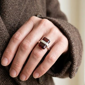 May include: A silver ring with a rectangular, deep red gemstone. The gemstone is set with a horizontal silver band. The ring is worn on a finger, with a brown jacket in the background.