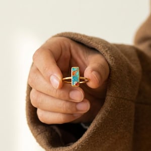May include: A gold-colored ring with a rectangular turquoise stone featuring orange and red accents. The ring is held in a hand, showcasing its design and color. The ring is a piece of jewelry.