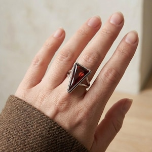 May include: A silver ring with a large, triangular, deep red gemstone. The gemstone is set in a silver frame with a dotted border. The ring has a unique, crossed band design, creating a bold and eye-catching piece of jewellery.