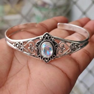 May include: A silver cuff bracelet with an oval, iridescent gemstone. The bracelet has detailed scrollwork and a decorative setting around the gemstone. The open design allows for easy wear, with a circumference of approximately 17 cm.