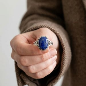 May include: A silver ring featuring a large, oval-shaped, deep blue lapis lazuli gemstone with subtle gold-colored flecks. The metal band has a split design that tapers toward the stone, accented by small, raised silver spheres on each side.