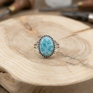 May include: Oval-shaped sterling silver ring featuring a light blue larimar gemstone. The ring has a beaded border around the stone and split shank design. The ring is set on a wooden surface.