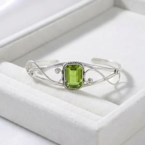 May include: A silver cuff bracelet featuring a central rectangular, emerald-cut light green gemstone. The metal band displays a delicate, openwork scroll design with twisted rope detailing surrounding the stone setting and small silver spheres.