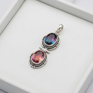 Puede incluir: Colgante de plata con dos piedras preciosas ovales. La piedra superior presenta un degradado de púrpura y azul, la inferior una mezcla de rosa y naranja. El colgante tiene un borde detallado con cuentas y un lazo para una cadena.
