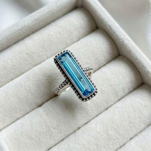 May include: A silver ring featuring a rectangular, light blue gemstone. The gemstone is framed by a beaded edge. The ring is displayed on a white surface.