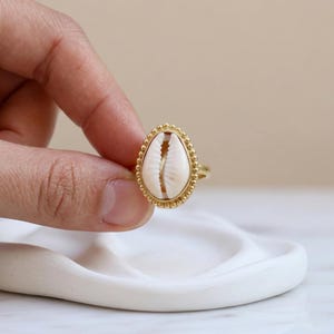 May include: A gold-colored ring featuring an oval-shaped, cream-colored seashell. The shell is set within a beaded gold frame. The ring is held up by a hand, showcasing the ring's design and details. The ring is a piece of jewelry.