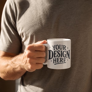 以下が含まれることがあります： 「YOUR DESIGN HERE」の文字が濃い青色で書かれた白いセラミックマグカップ。グレーのTシャツを着た人が持っています。マグカップには標準的なハンドルが付いています。