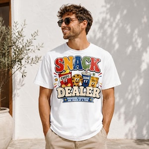 May include: White t-shirt with a colorful graphic that reads "SNACK DEALER" above cartoon images of chips, a cookie, soda, and treats. The text "PROVIDING HAPPINESS ONE SNACK AT A TIME" is below the word "DEALER".