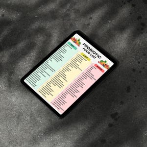 K&ouml;nnte beinhalten: Ein digitales Tablet zeigt eine "Probiotische Lebensmittelliste" mit den Abschnitten "Essen", "Einschr&auml;nken" und "Vermeiden". Die Liste enth&auml;lt verschiedene Lebensmittel, mit farbenfrohen Grafiken von Obst und Gem&uuml;se. Das Tablet hat einen schwarzen Rahmen und liegt auf einem dunkelgrauen Hintergrund.