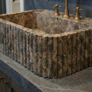 Può includere: Un lavabo rettangolare in marmo marrone con un esterno strutturato e a coste verticali. Il lavabo ha un rubinetto integrato e due maniglie con finitura dorata. Il lavabo è appoggiato su un piano di lavoro grigio scuro.