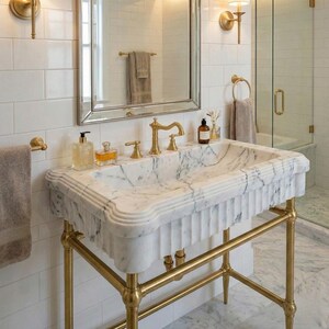 Puede incluir: Un lavabo de baño de mármol con grifos dorados y un soporte dorado. El lavabo tiene un borde decorativo y una cubeta rectangular. Un espejo y apliques de pared son visibles en el fondo.